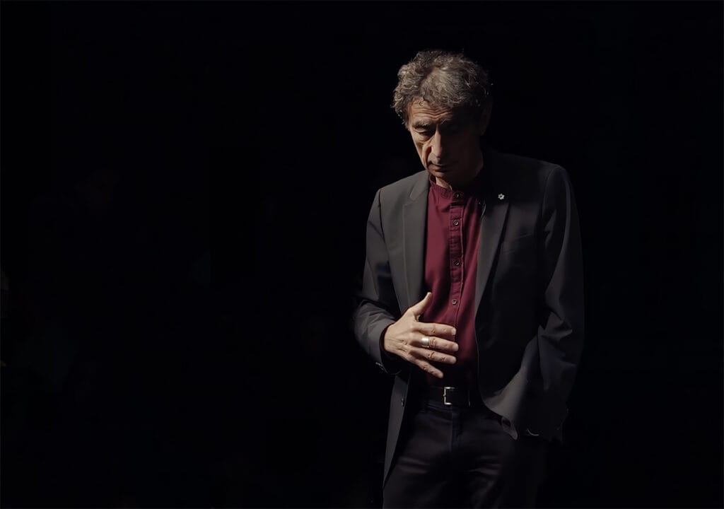 Dr. Gabor Mate is standing in a semi-darl soft light, giving a lecture