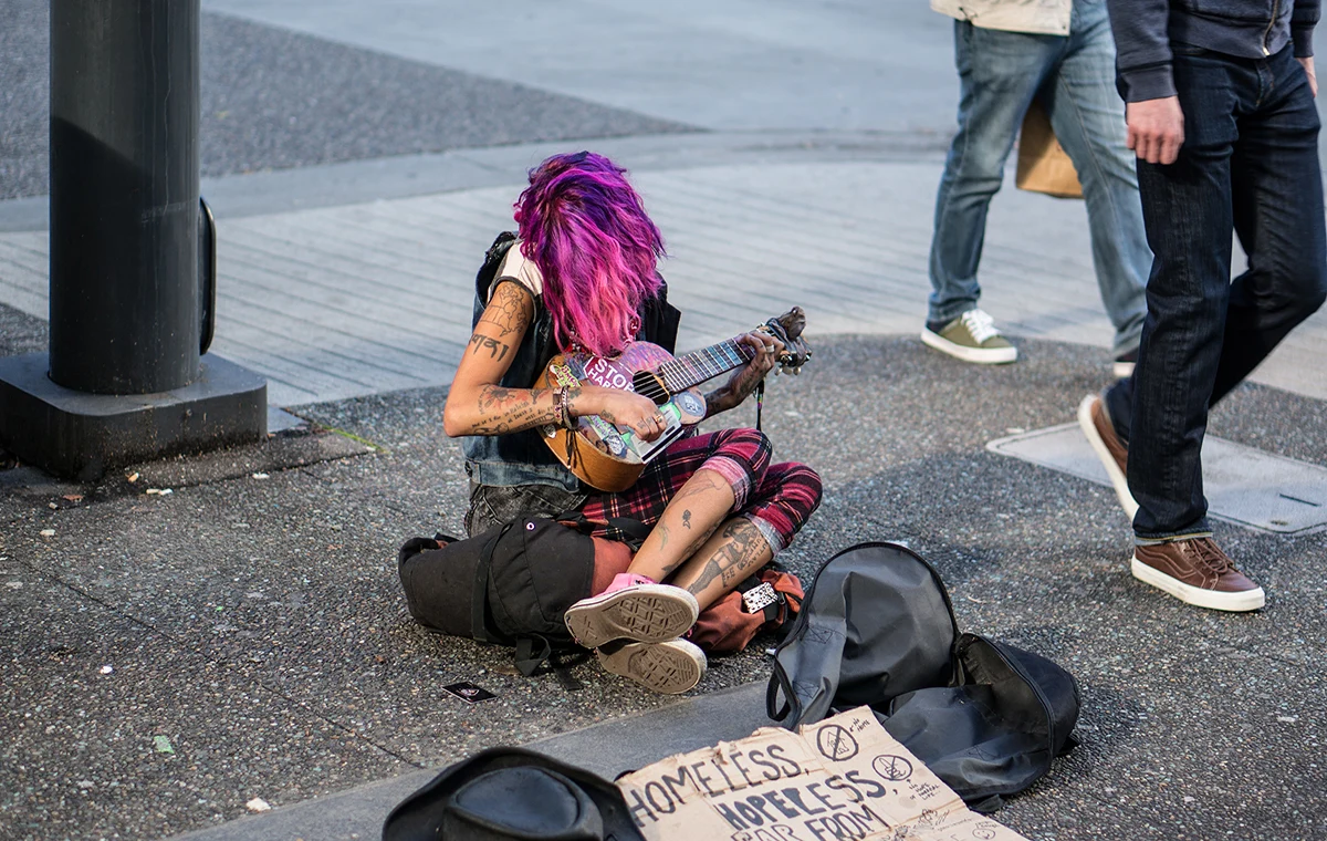 A picture of a homeless woman on a streetside playing a guitar with a sign in front stating that she is homeless