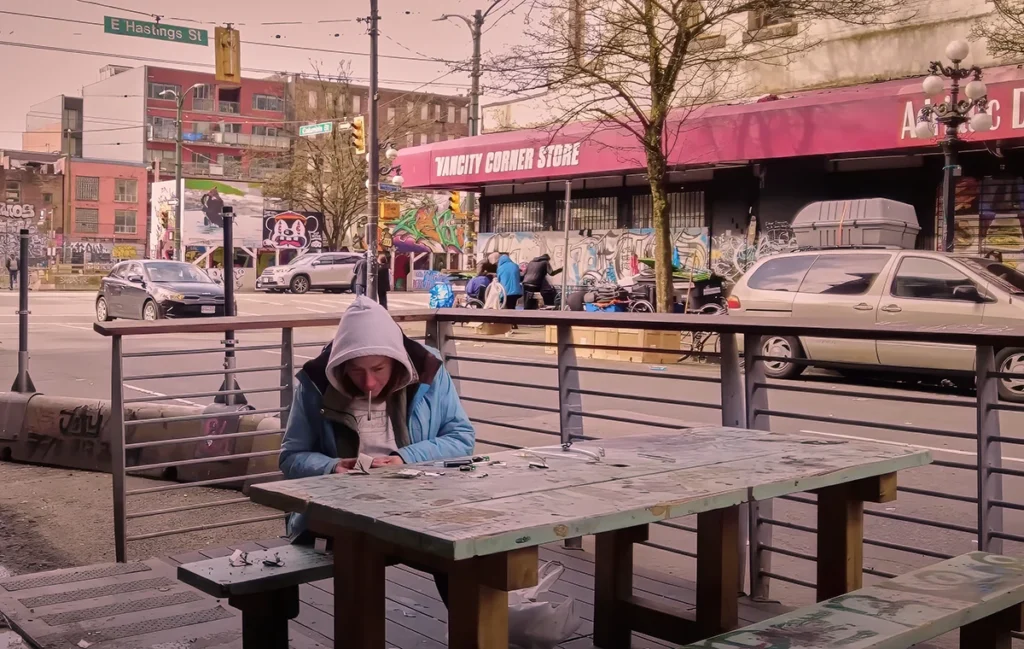 An image of a homeless person sitting on a bench consuming substances in the middle of a busy urban environment