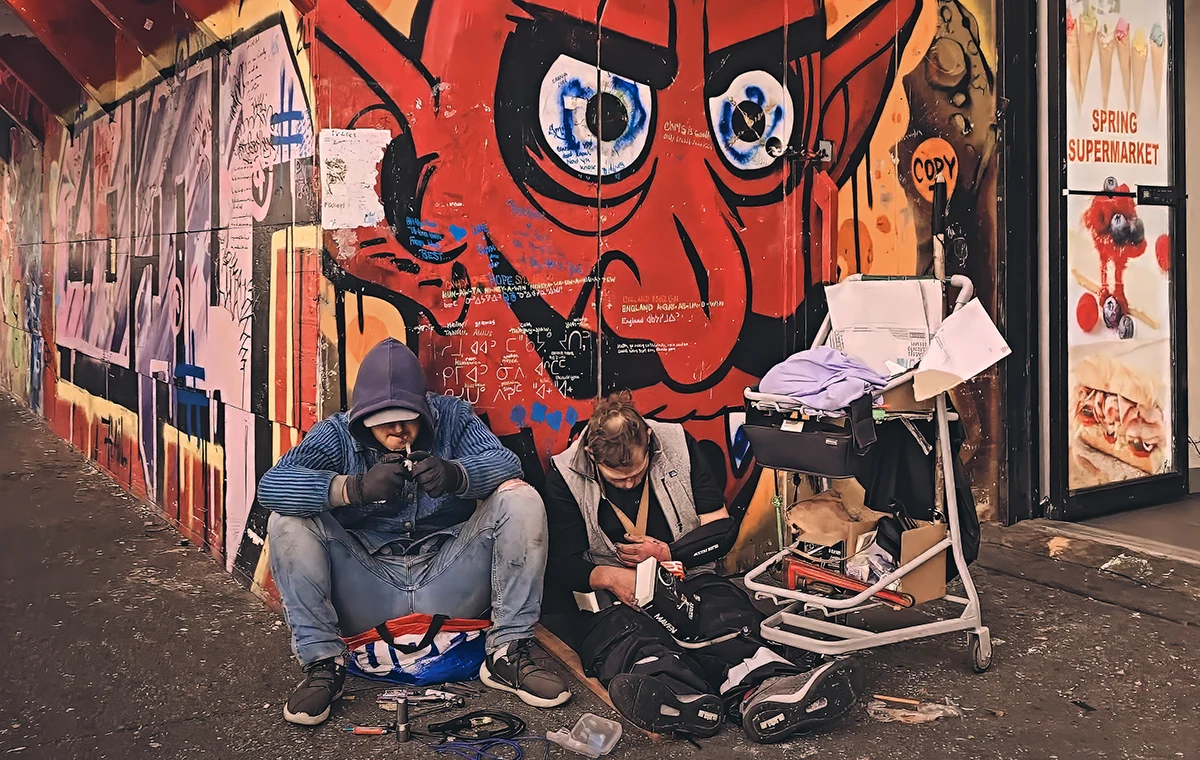 Two men pictured on the sidewalk in visible distress leaning against the wall of graffiti