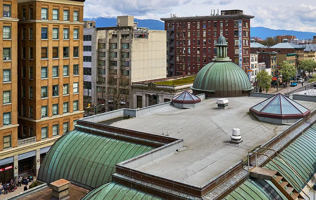 A view from Carnegie Library overlooking East Hastings Street in Downtown Eastside Vancouver, showing a bustling urban scene with people walking along the sidewalks and traffic passing by. The image captures the historic buildings and the lively, dynamic neighbourhood outside.