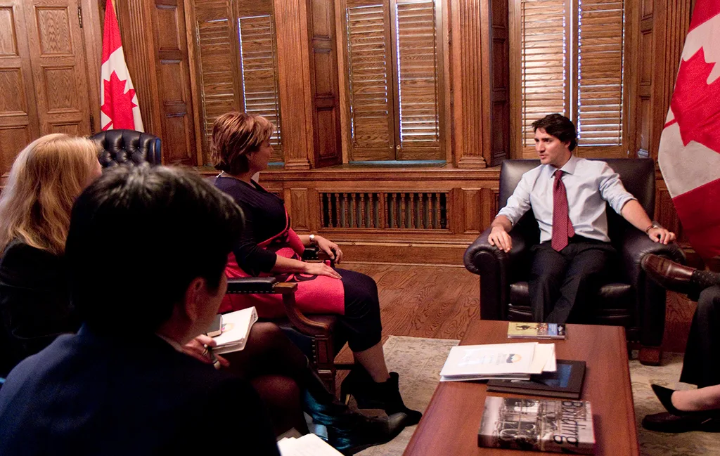 Canadian Prime Minister Justin Trudeau sits in discussion with a group of officials in a formal meeting room, flanked by Canadian flags and a wooden interior.