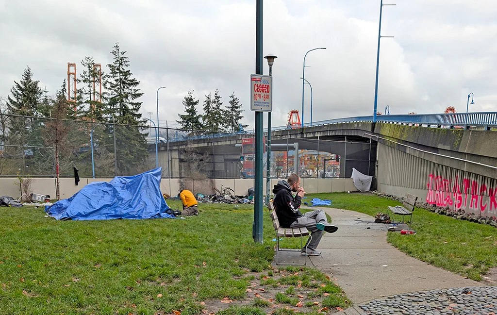 An encampment in a Vancouver park with a blue tarp shelter, a man sitting on a bench, and a sign indicating park closure hours, highlighting homelessness issues.