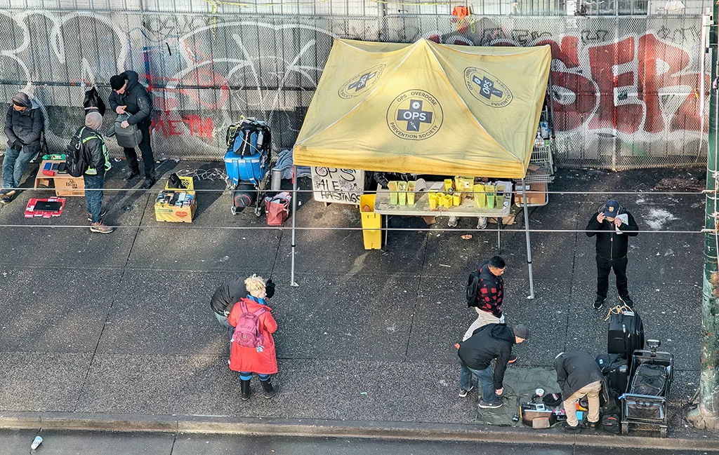 An overhead view of a supervised drug consumption site in an urban setting, featuring a yellow tent labeled 'OPS - Overdose Prevention Society,' surrounded by individuals engaging with staff and accessing services, against a backdrop of graffiti-covered walls.