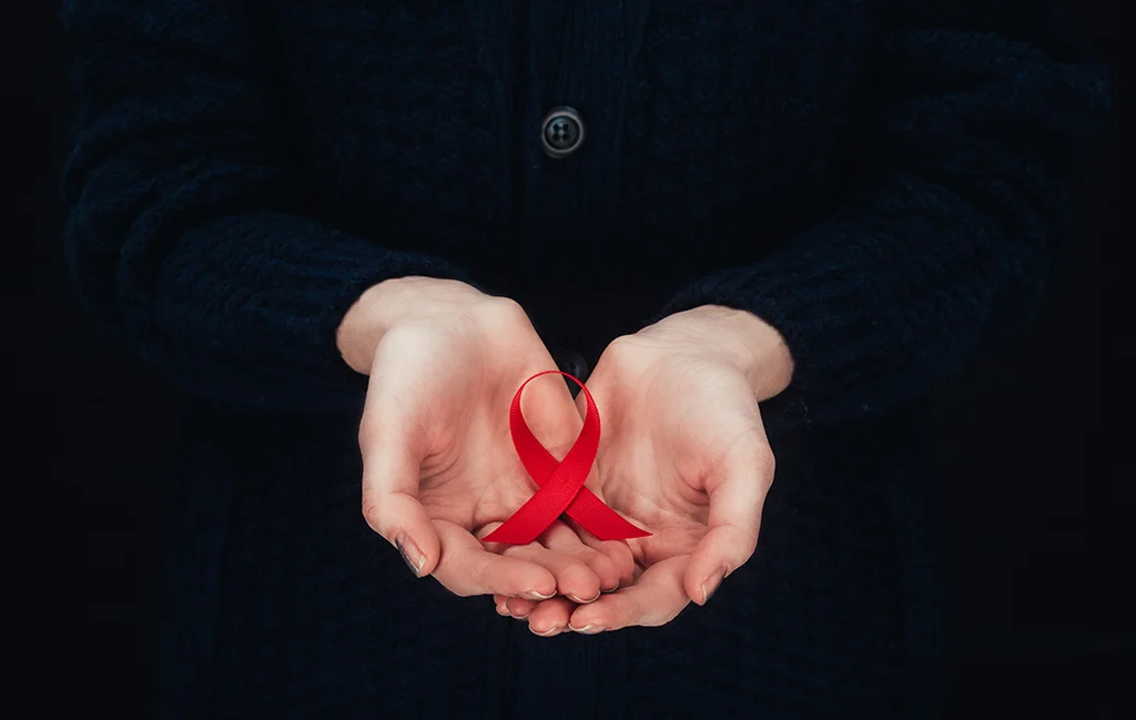 A person wearing a dark coat holds a red ribbon in their cupped hands, symbolizing HIV/AIDS awareness and the ongoing fight against the epidemic.
