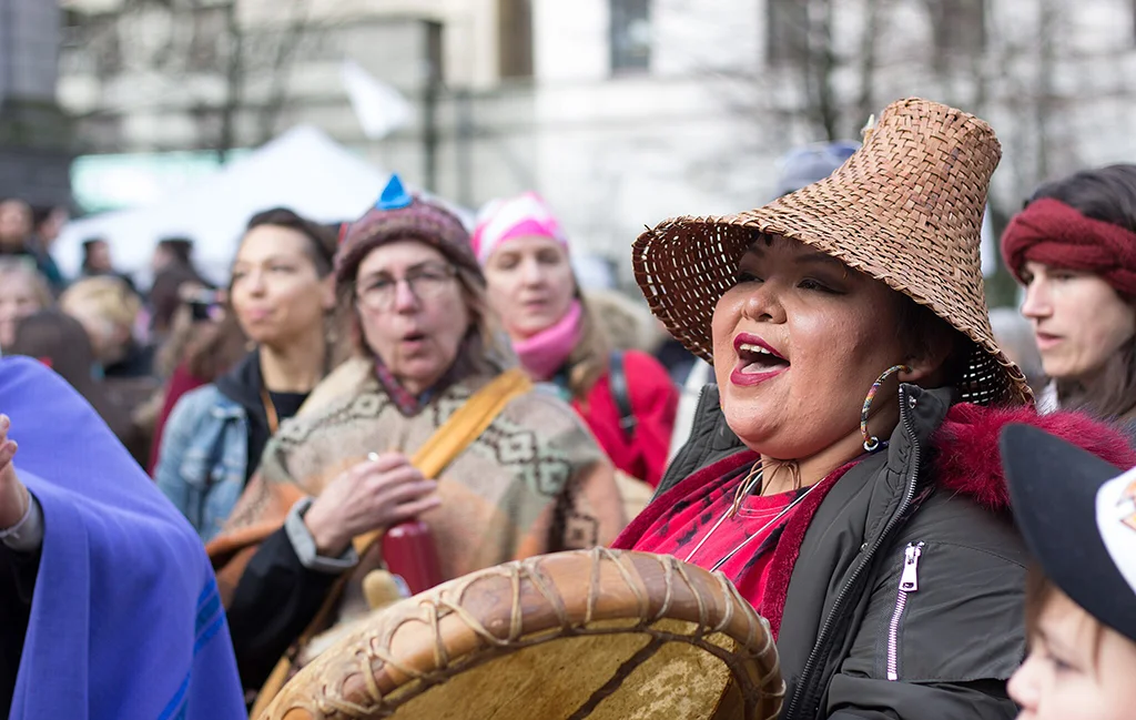 A group of women participate in the Women’s Memorial March, with one woman wearing a woven cedar hat and holding a drum, singing during the gathering.