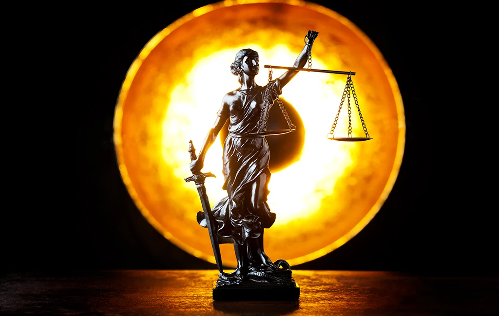 A statue of Lady Justice holding scales and a sword, silhouetted against a bright glowing circular background, symbolizing legal fairness and the complexities of the justice system.