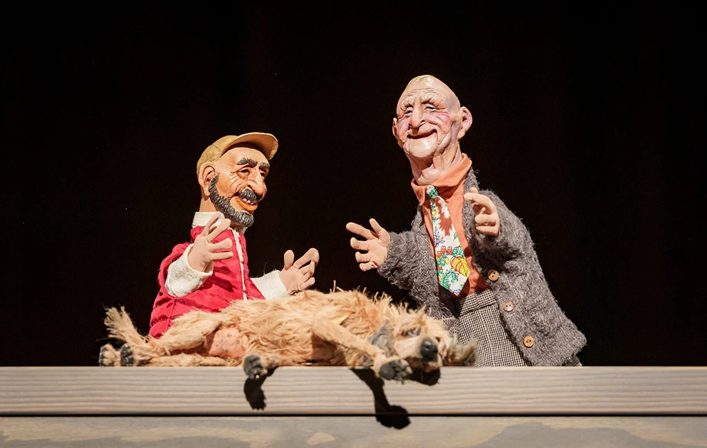 Two marionette puppets from Wonderful Joe are on stage, with one resembling an elderly man wearing a floral tie and cardigan, and the other a bearded man in a red jacket, alongside a puppet dog lying on a table.