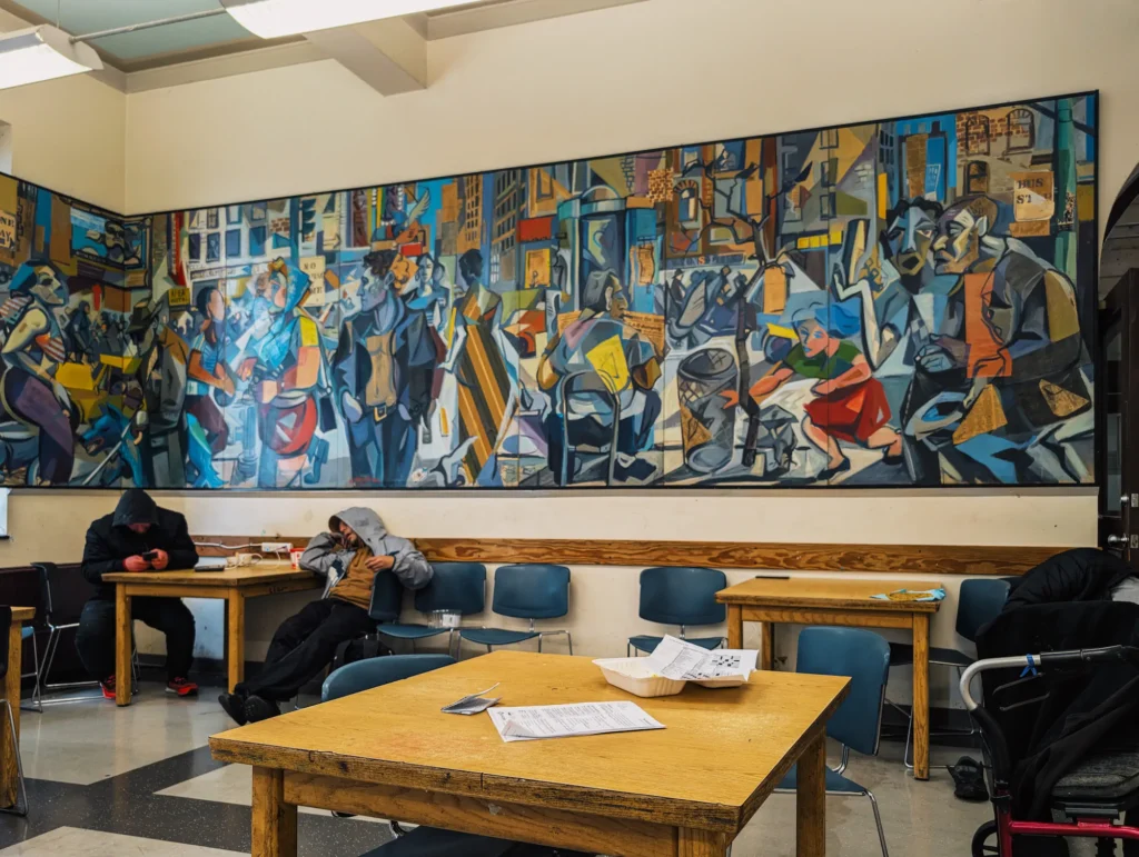 Wide view of Richard Tetrault's large six-by-24-foot mural mounted on an interior wall inside Carnegie Community Centre above tables and chairs.