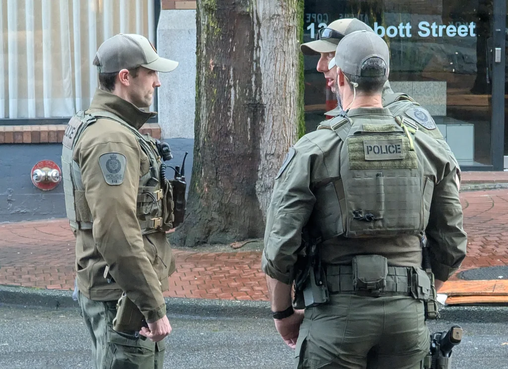 Three police officers wearing tactical uniforms and protective vests standing together on a city sidewalk on Abbott Street in Vancouver.