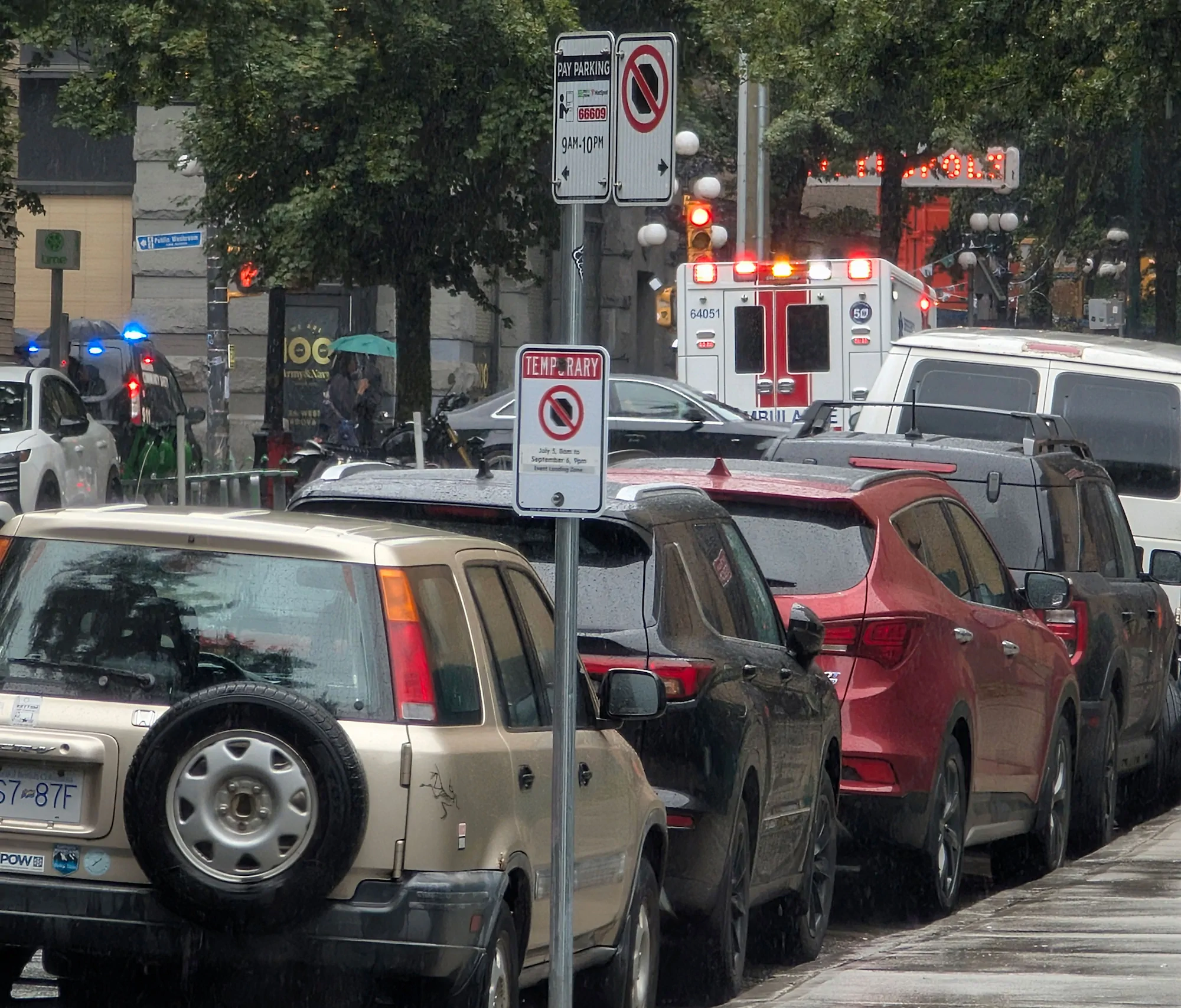 A rainy city street scene featuring a line of closely parked cars in the foreground, including a tan SUV with a rear-mounted spare tire parked next to temporary "No Parking" signs. In the background intersection, a white ambulance is stopped with its red emergency lights actively flashing, while the blue and red lights of a police vehicle are visible further back on the left side of the street. Trees and a pedestrian with a green umbrella line the wet sidewalk near a red traffic light.