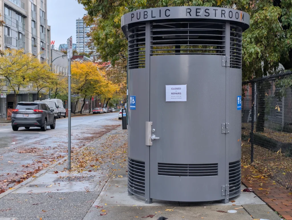 A grey cylindrical Portland Loo public restroom with a "Closed for Repairs" sign posted on the door on a leaf-strewn Vancouver sidewalk.