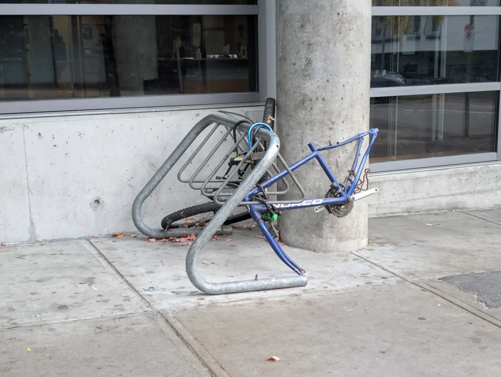 A stripped blue bicycle frame missing its wheels and handlebars, U-locked to a metal bike rack on a concrete sidewalk.