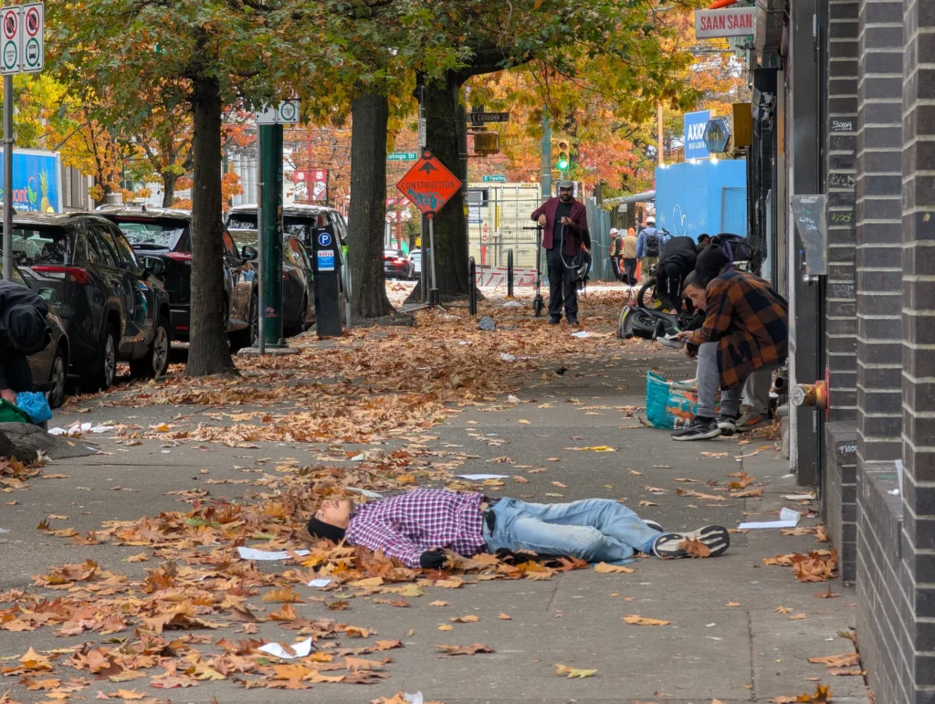 A sidewalk covered in fallen autumn leaves on East Hastings Street in Vancouver with an individual lying on the ground near local storefronts and passersby.
