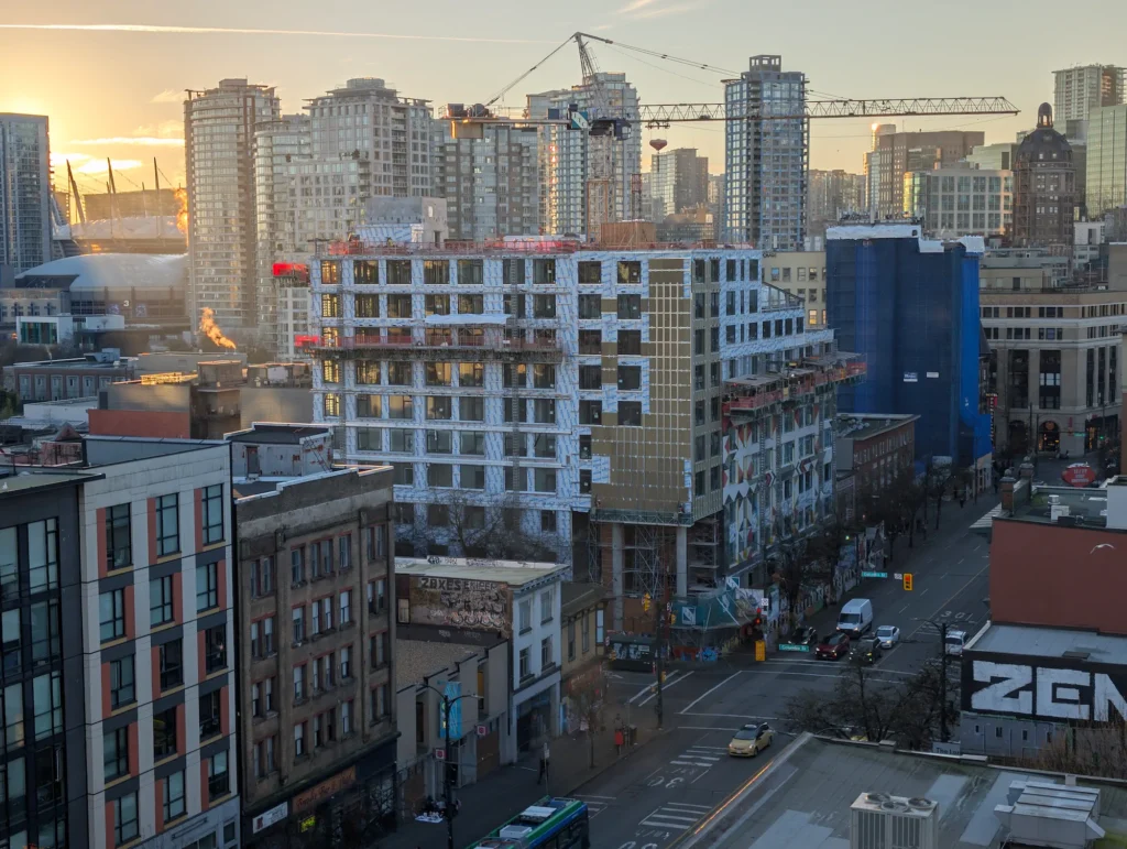 An elevated view of a multi-story building under construction with a tower crane overhead on East Hastings Street, with the Vancouver downtown skyline in the background.