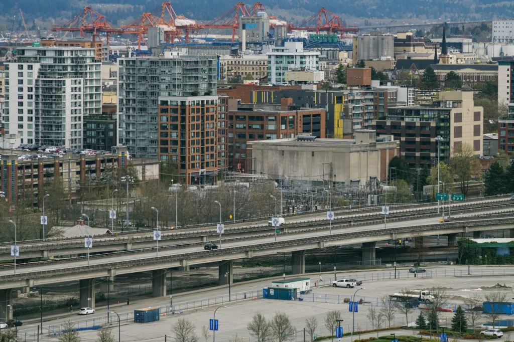 An elevated daytime view of the twin concrete Georgia and Dunsmuir viaducts spanning horizontally across the frame with sparse traffic. Below the elevated roadways are empty, fenced-off dirt lots and paved parking areas. The background features a dense mix of urban mid-rise buildings, an electrical substation, and large orange shipping cranes with mountains visible in the far distance.